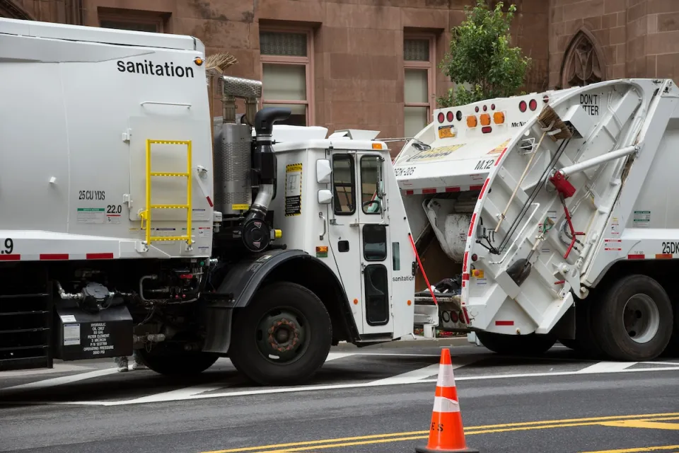 Two white garbage trucks stopped on New York City street.
